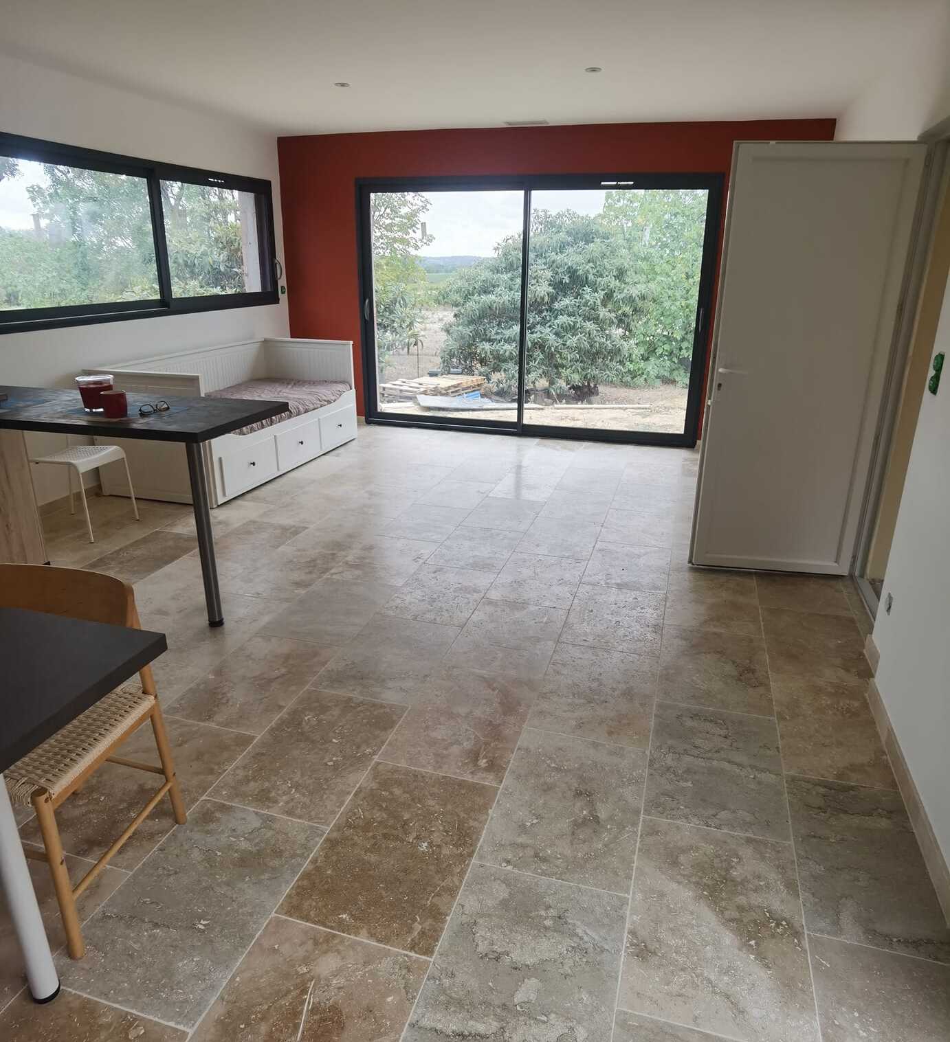 Pièce de vie moderne : sol en travertin beige, mur terracotta, baie vitrée sur jardin, lit d'appoint et table en bois.