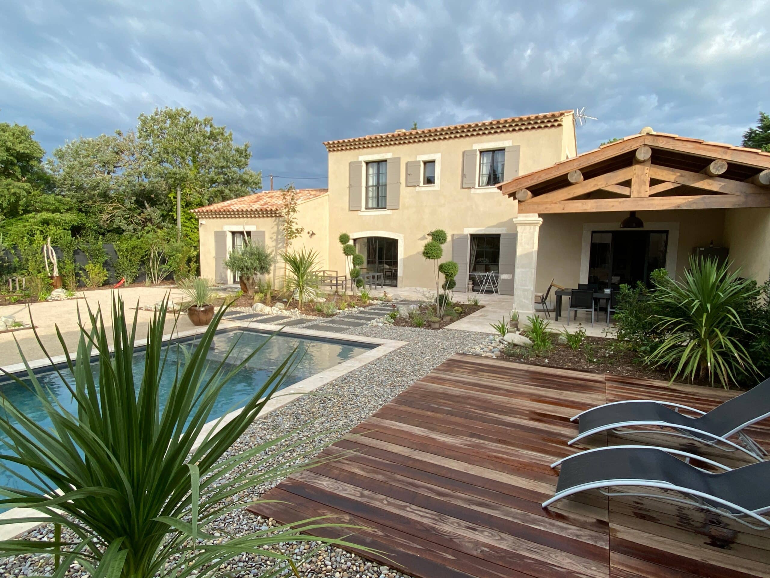 Maison provençale avec piscine moderne, terrasse en bois et jardin méditerranéen sous un ciel nuageux.