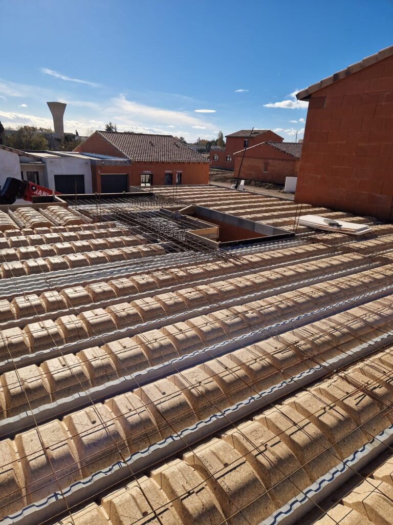 Plancher de toiture en construction, avec hourdis isolants et ferraillage en attente du béton. Maisons Terreal et château d'eau sous ciel bleu.