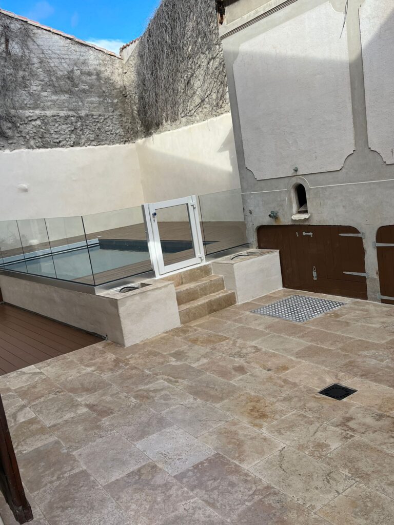 Cour intérieure: piscine et sol en travertin beige Cour pavée de travertin beige. Petit bassin moderne surélevé avec terrasse bois et barrière en verre. Murs anciens et portes en bois.