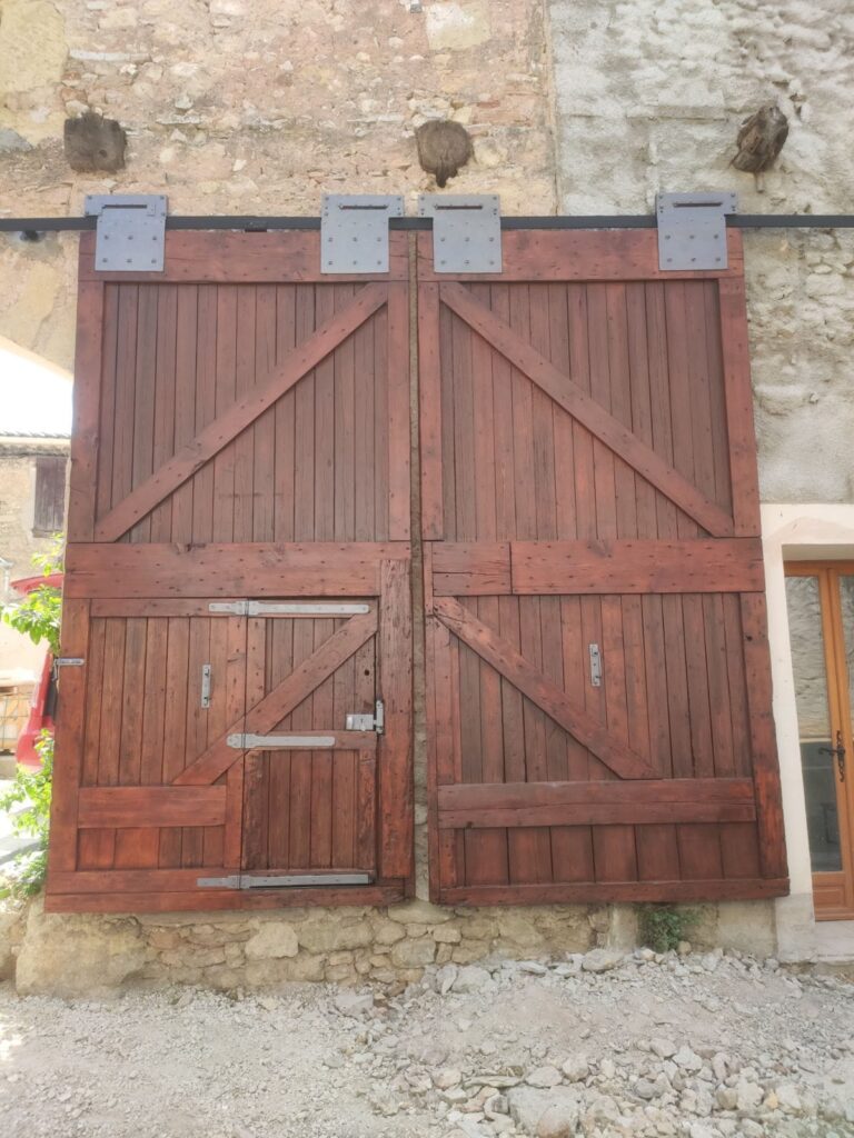 Porte de grange coulissante bois: charme et robustesse. Grande porte coulissante rustique en bois teinté rouge, style grange, avec ferronnerie métallique moderne sur un ancien mur de pierres.