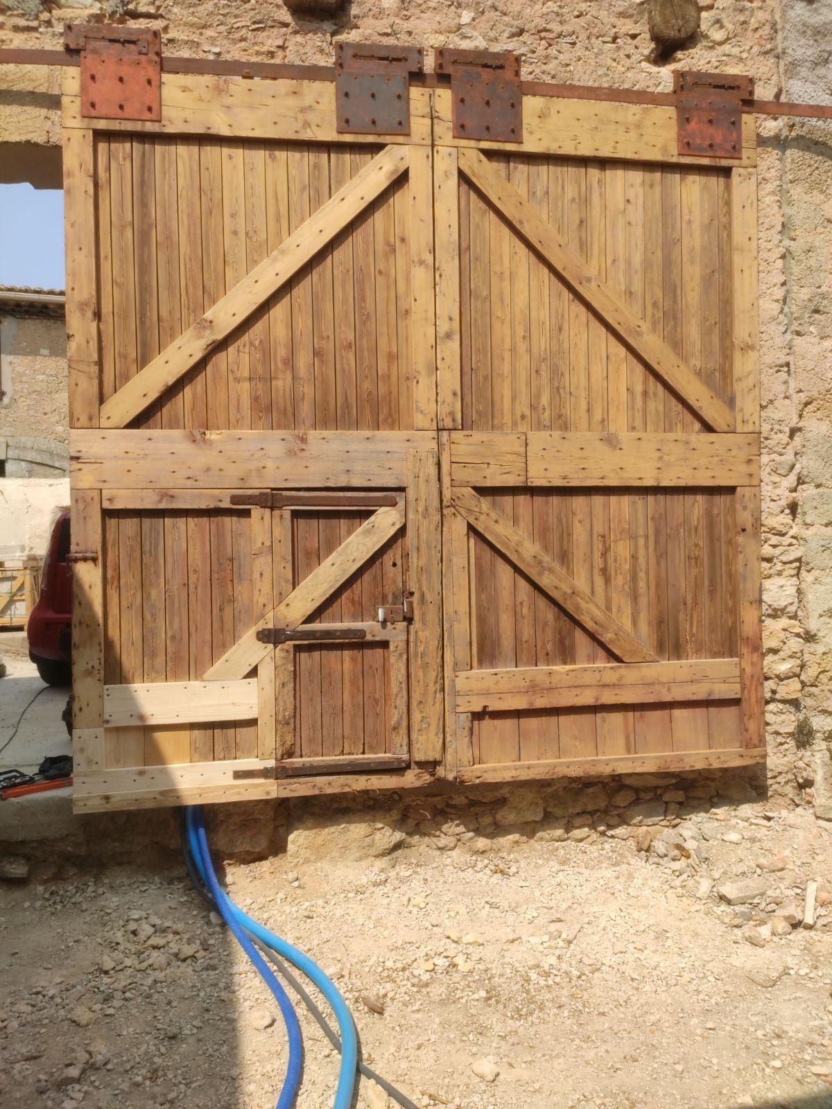 Porte coulissante de grange rustique en bois clair, renforcée de traverses diagonales. Porte piétonne dans le panneau gauche. Installée sur mur de pierre.