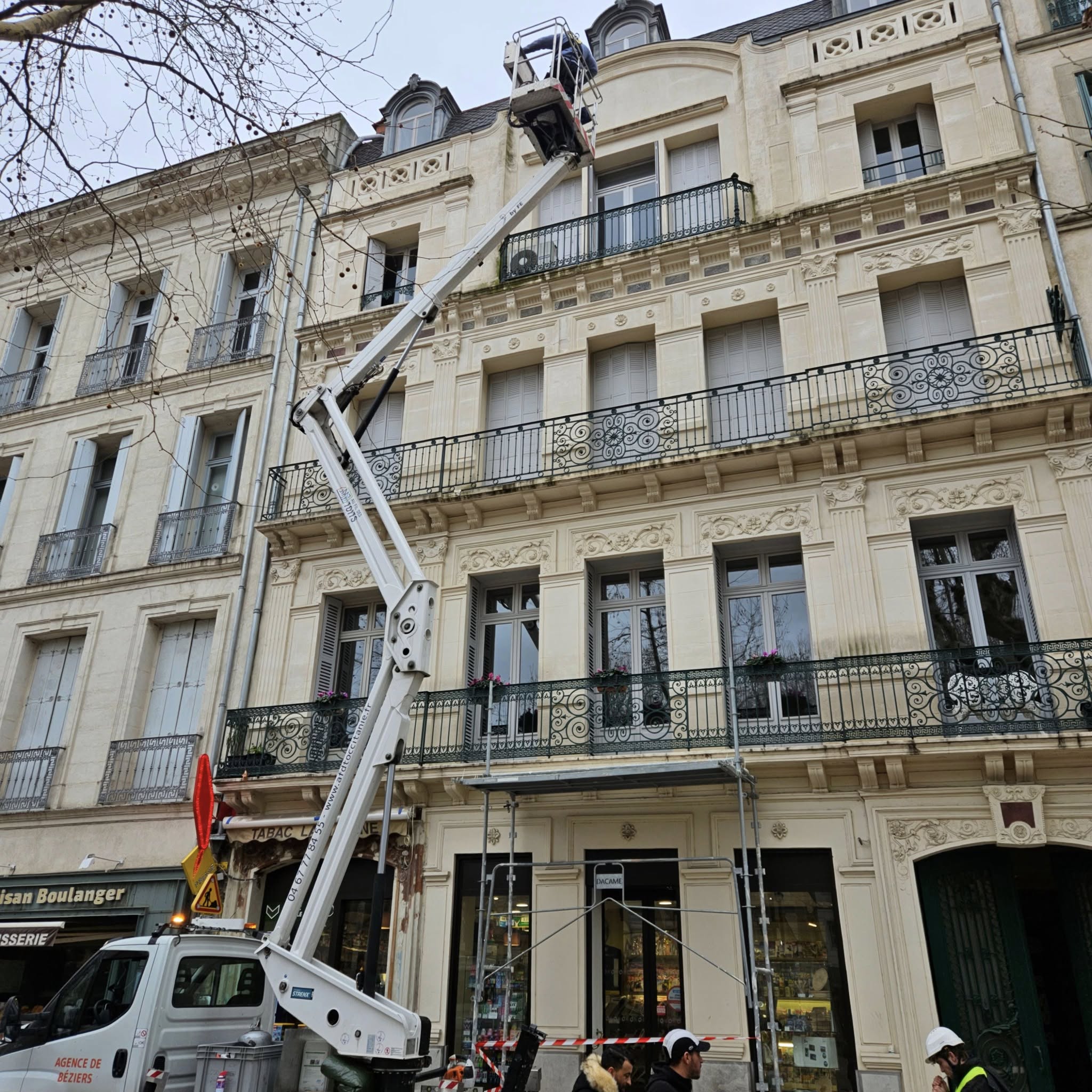 Rénovation d'une façade en pierre sculptée avec balcons en fer forgé à Béziers. Un ouvrier utilise une nacelle élévatrice.