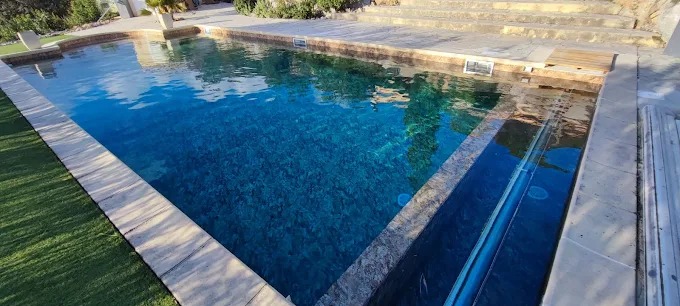 Piscine rectangulaire au carrelage bleu foncé avec plage en pierre naturelle et volet roulant immergé.