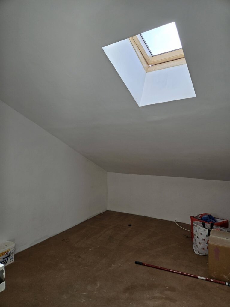 Chambre mansardée blanche avec fenêtre de toit Velux et sol brun en cours de rénovation.
