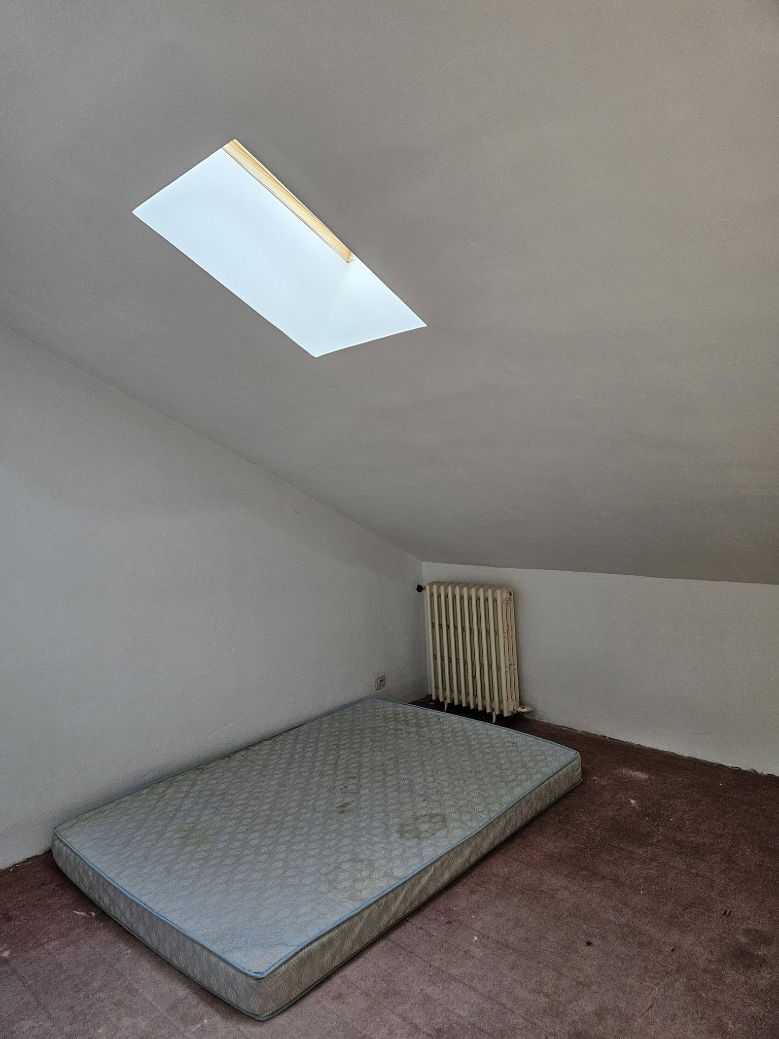 Chambre mansardée avec matelas au sol, vieux radiateur et fenêtre de toit.