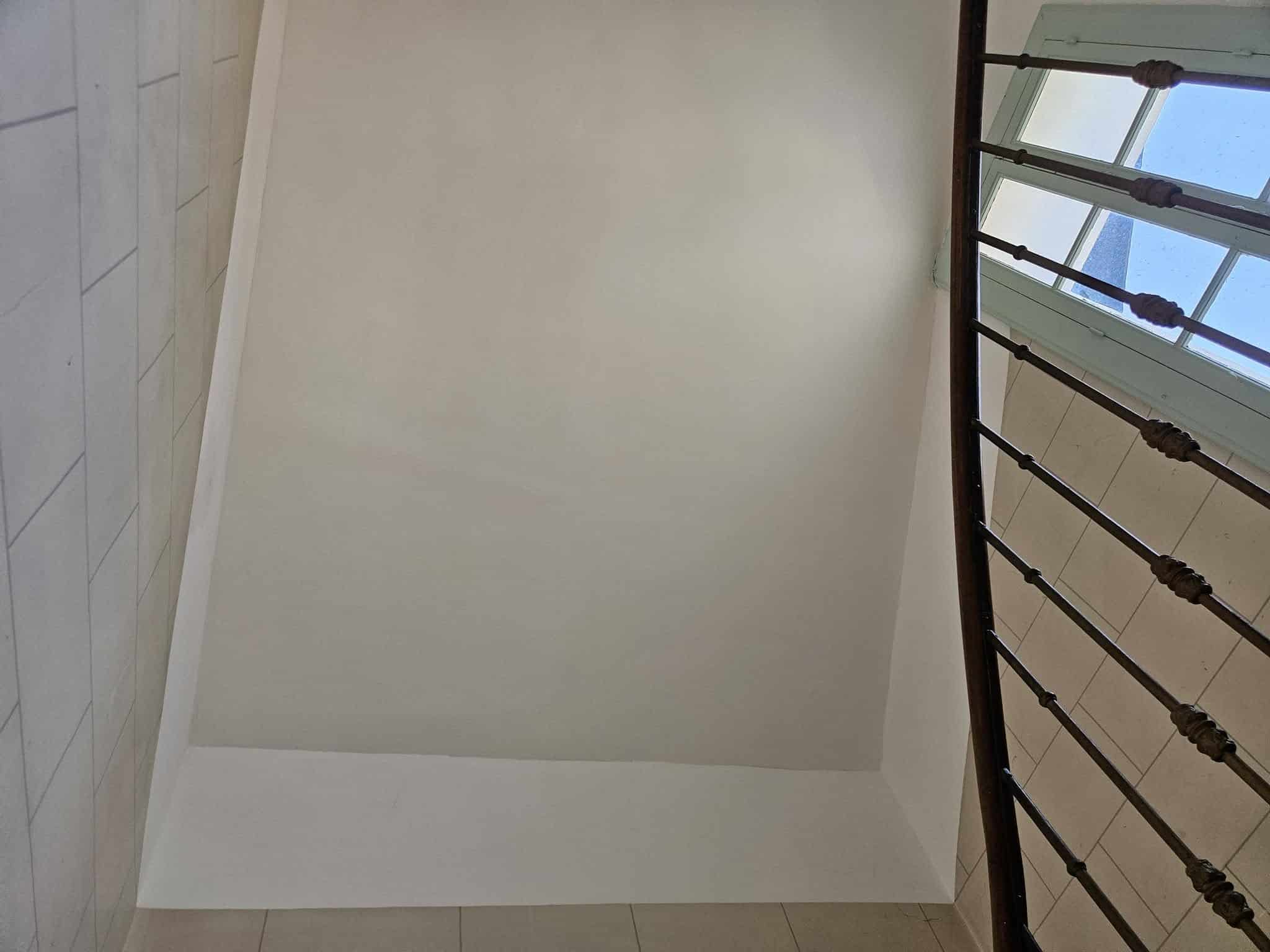 Escalier en colimaçon, rampe en bois sombre et murs blancs sous une fenêtre éclairée.