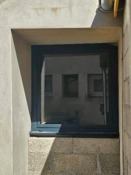 Fenêtre anthracite encastrée dans un mur en crépi avec soubassement en parpaings apparents.