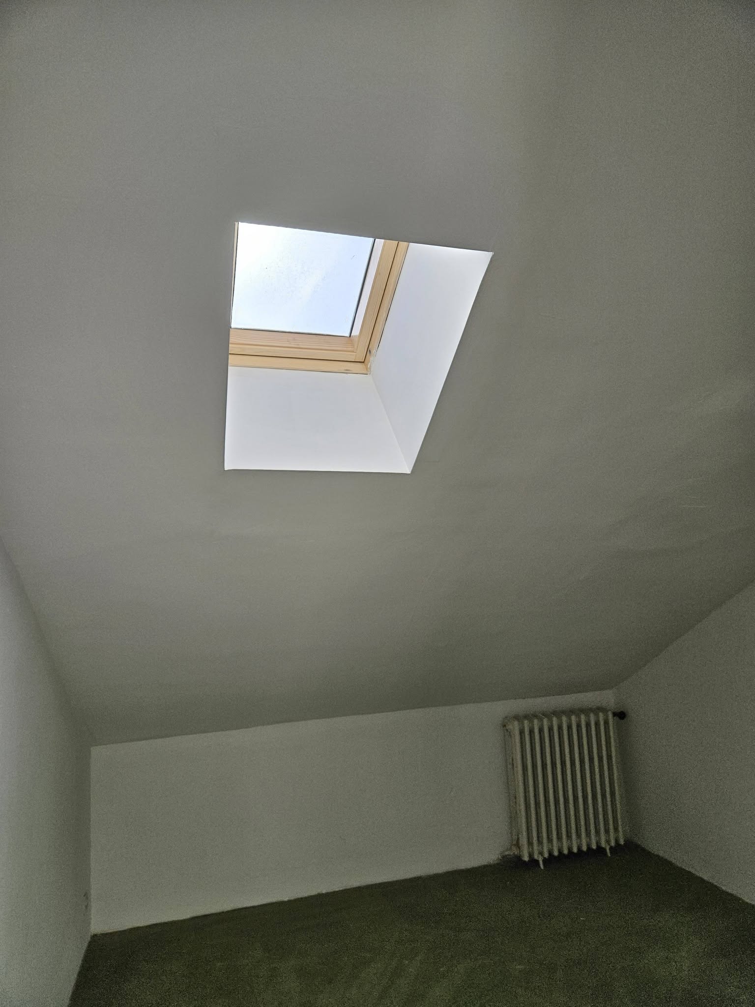 Fenêtre de toit Velux dans une chambre mansardée blanche avec radiateur et moquette verte.