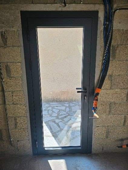 Porte Vitrée Cadre Anthracite sur Mur en Parpaings Porte vitrée à cadre gris anthracite installée dans un mur en parpaings bruts, vue sur patio pavé.