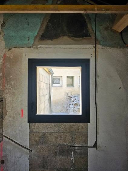 Fenêtre noire moderne installée sur un mur brut en rénovation, vue sur une cour intérieure.